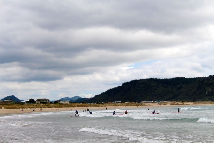 Surfers riding waves at scenic Whangamata Beach, under a cloudy sky with distant hills.