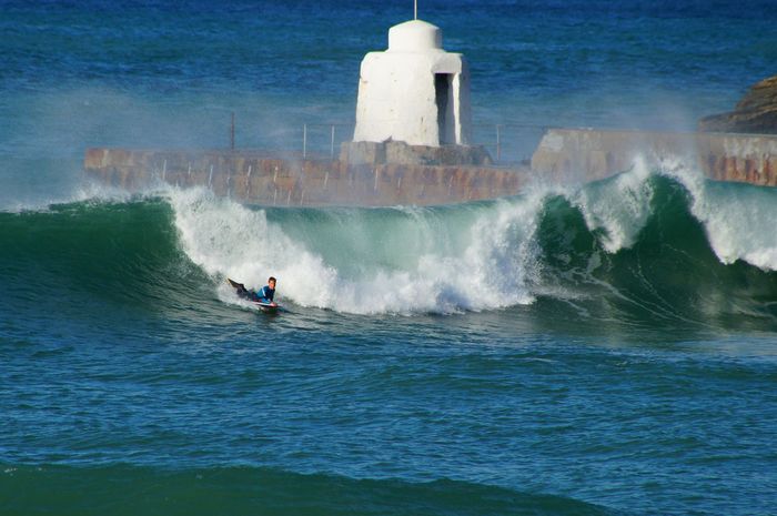 Surfer riding the waves with lighthouse in the background at Portreath, England.