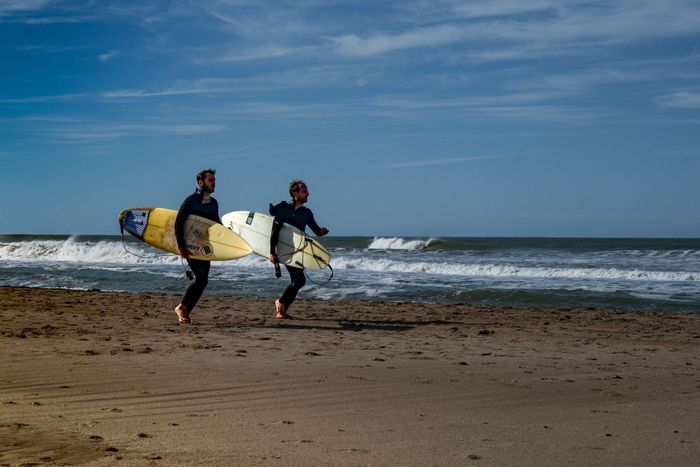 Two surfers with boards walk along Cariló beach in Argentina under a clear blue sky.