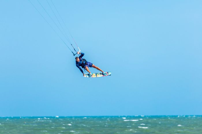 Kitesurfer performing an aerial trick against a clear blue sky over the ocean waves.