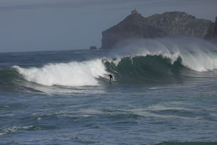 Dynamic shot of a surfer navigating large waves near a rocky Basque coastline.