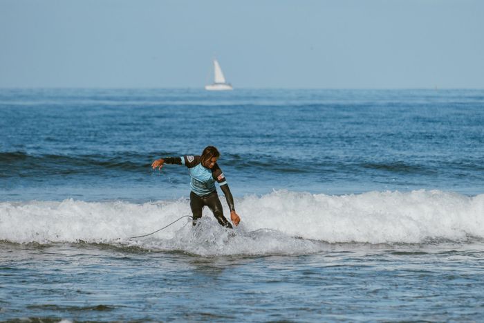 Surfer in wetsuit conquering a wave, with a sailboat on the horizon.