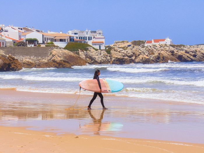 A surfer carrying a colorful surfboard strolls along the scenic beach in Peniche, Portugal.