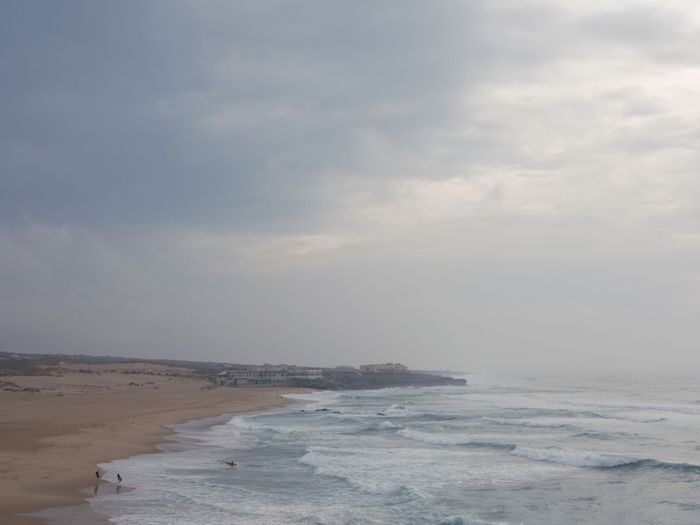 A tranquil beach scene under an overcast sky with gentle waves and distant surfers.