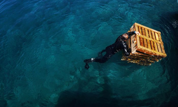 From above back view of unrecognizable male in diving suit swimming in rippled sea with wooden box