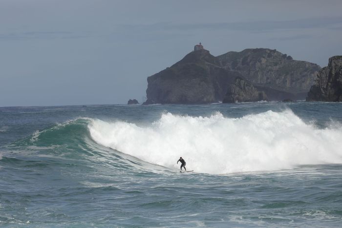 A surfer maneuvers through large waves with a rocky coastline in the background in Bakio, Spain.