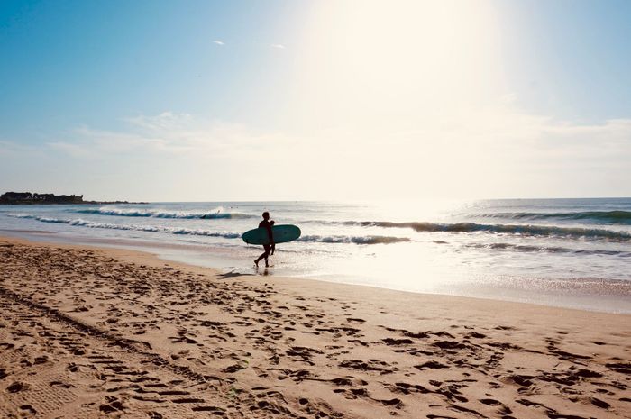A lone surfer walks along Matosinhos Beach in Portugal, carrying a surfboard during a beautiful sunset.