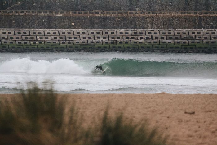 A surfer catches a wave near a concrete breakwater on a cloudy day, blending leisure and nature.