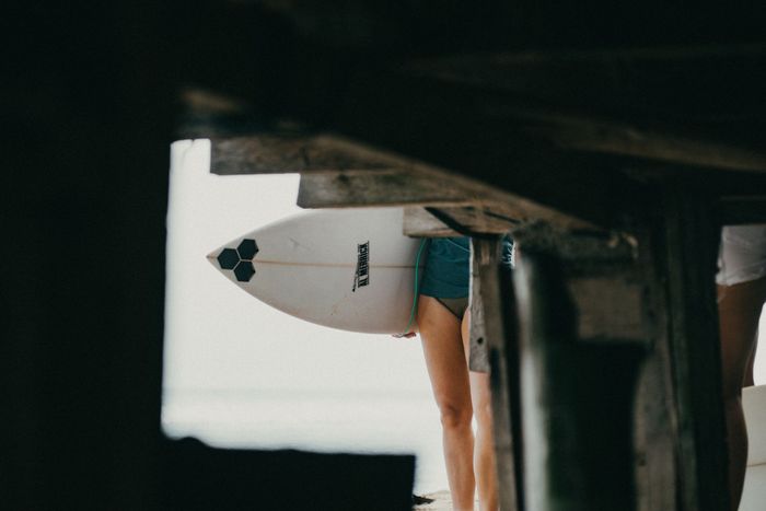 Surfer with board under a pier in Bali, creating a unique perspective on beach life.