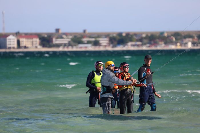 A group of people receiving a kiteboarding lesson in shallow sea waters, equipped with helmets and gear.