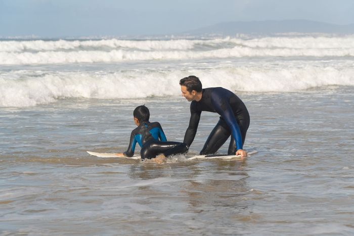 A father teaching his son how to surf on a Lisbon beach. The waves crash as they bond over this water sport.