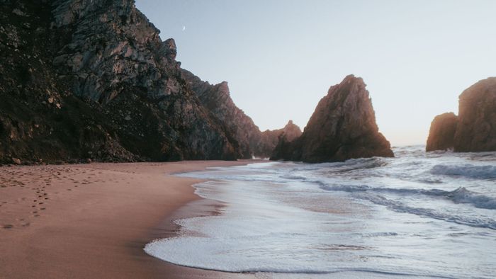 Breathtaking view of Praia da Ursa beach's rocky coastline during sunrise with gentle waves and soft sand.