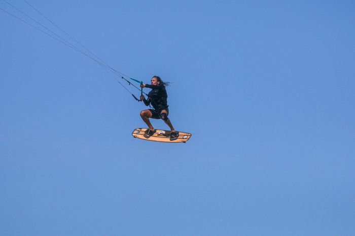 A kitesurfer performs an aerial trick in the sky, showcasing skill and action against a clear blue backdrop.