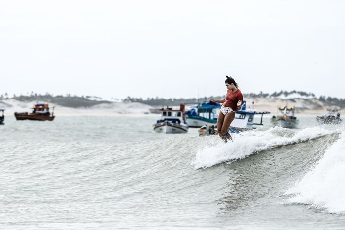 Woman surfing on ocean wave with boats in background at Baía Formosa, Brazil.