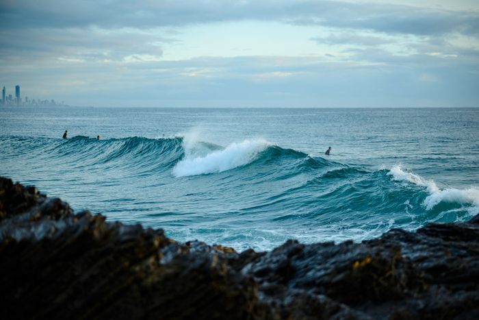 Surfers catching waves on Currumbin Beach with rocky shoreline and distant city skyline.