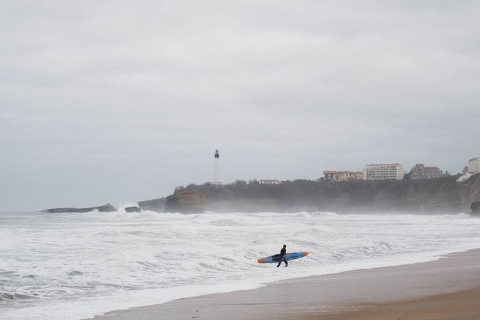 Surfer carrying a board on Biarritz beach with lighthouse and ocean waves in view. Perfect for travel and lifestyle themes.