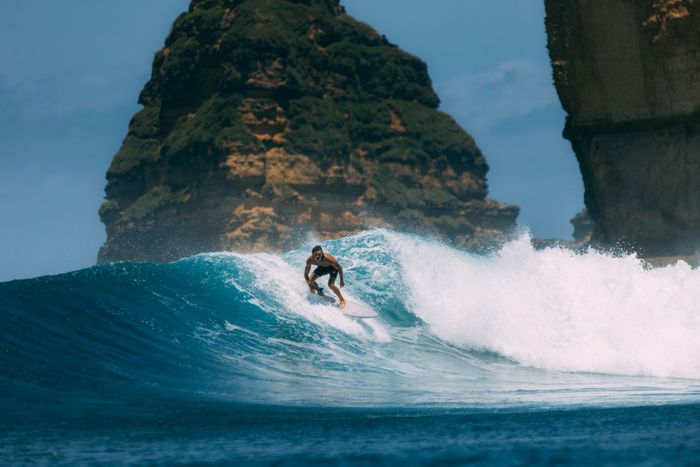 A skilled surfer catching a wave in Indonesia, with a dramatic rocky coastline backdrop.