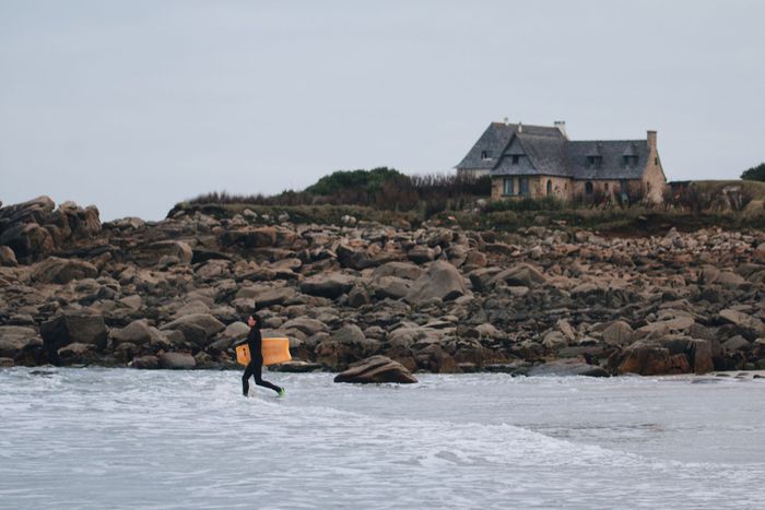Surfer carrying a bodyboard towards ocean waves near a stone house on a rocky shoreline.