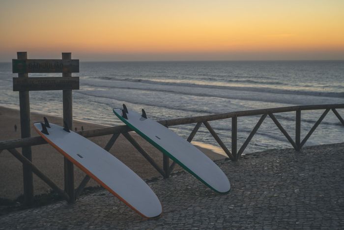 A serene view of surfboards on a wooden fence overlooking Ericeira beach at sunset.