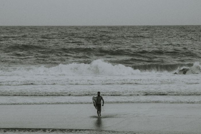 A lone surfer carrying a surfboard on a monochrome beach in Itacaré, Brazil.