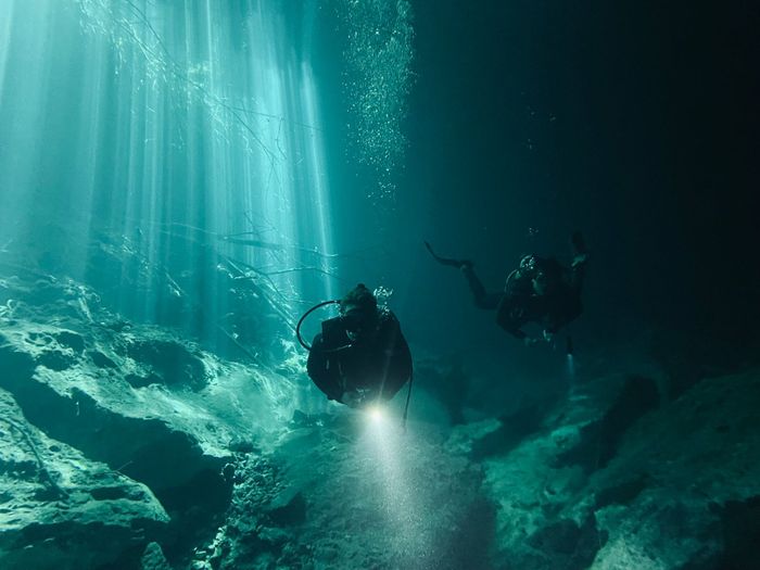 Scuba divers navigate underwater, exploring with flashlights and enjoying sunlight rays.