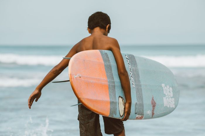 A young surfer walks on a Honolulu beach, carrying a surfboard on a sunny day.