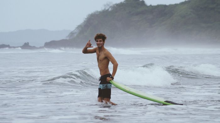 A young surfer stands confidently in ocean waves with his surfboard on a misty day.