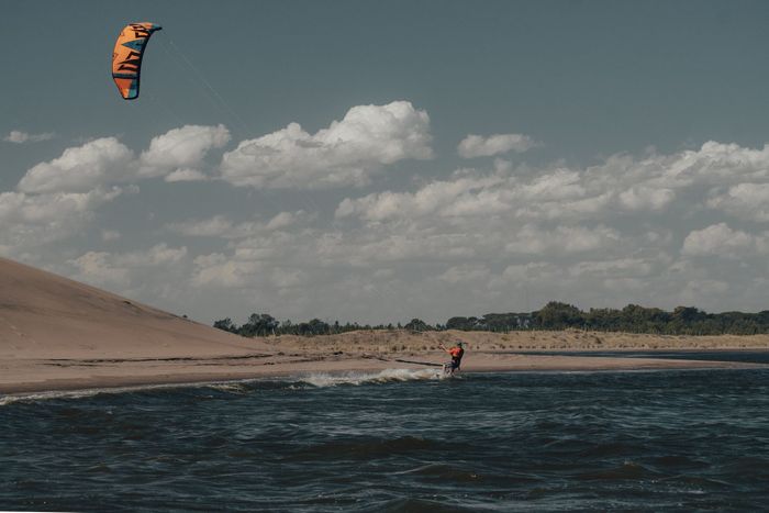 A kitesurfer expertly glides over the waves along a sandy coastline under a cloudy sky.