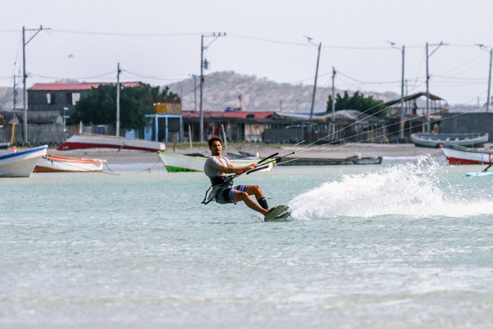 A man kitesurfing on a sunny day near a beach with colorful fishing boats in the background.