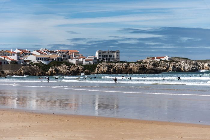 Surfers riding waves near a charming beach town with rocky shores.
