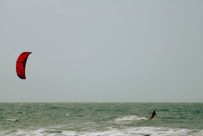 A lone kitesurfer glides across the ocean waves under a red kite on a clear day.