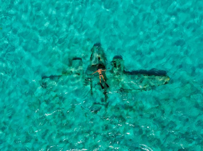 Snorkeler explores a submerged airplane wreck in clear turquoise waters from above.