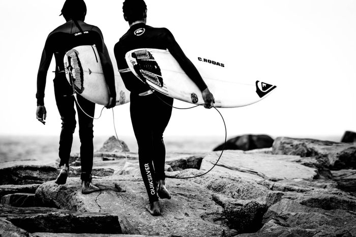 Two surfers walking on rocks with surfboards by the sea in black and white.