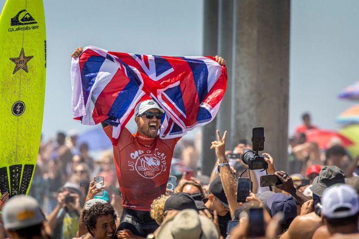 Surfer celebrates victory hoisting Union Jack flag, surrounded by a lively crowd.