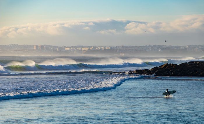 A surfer approaches ocean waves at Costa da Caparica beach with a clear sky backdrop.