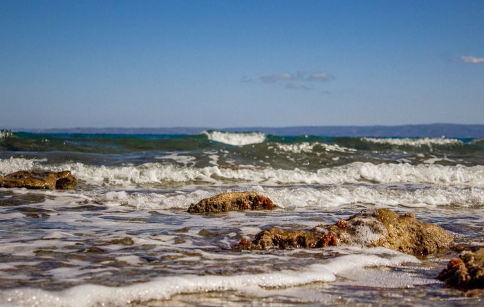 Waves gently splashing on the rocky shore in Hanioti, Greece under a clear blue sky.
