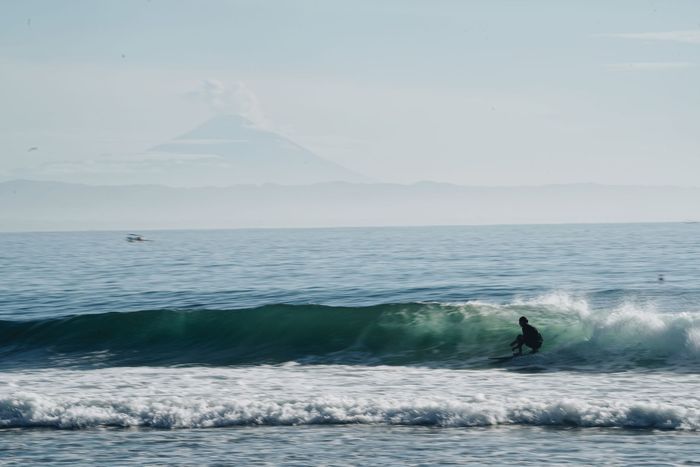 A surfer skillfully rides ocean waves with a stunning volcano backdrop, captured during daylight.