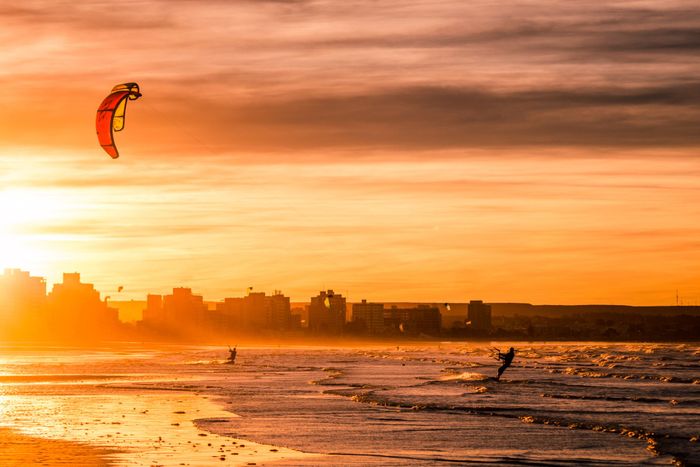 Kite surfers riding waves at sunset by Puerto Madryn's coastline. Vibrant sky and sea landscape.