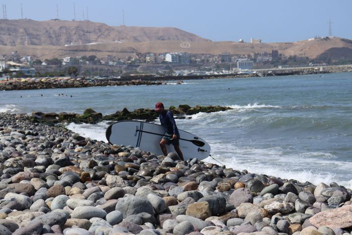 A surfer carrying a board along the rocky beach of Lima, Peru on a sunny day.